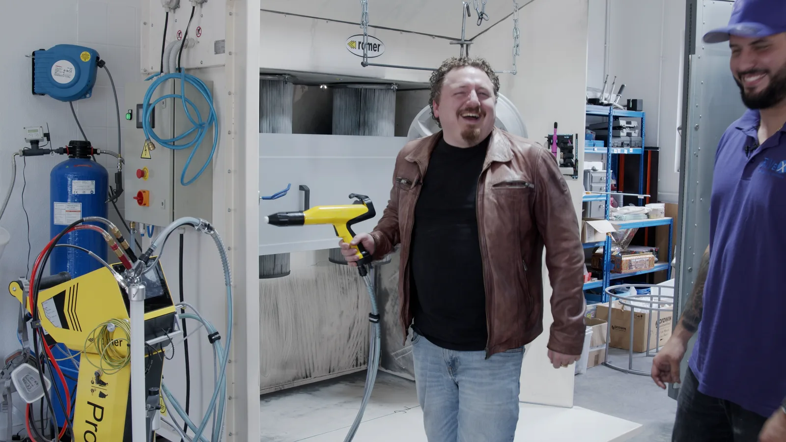 Ein Mann haelt eine gelbe Pulverbeschichtungspistole in der Hand, waehrend er vor einer Pulverbeschichtungskabine steht.