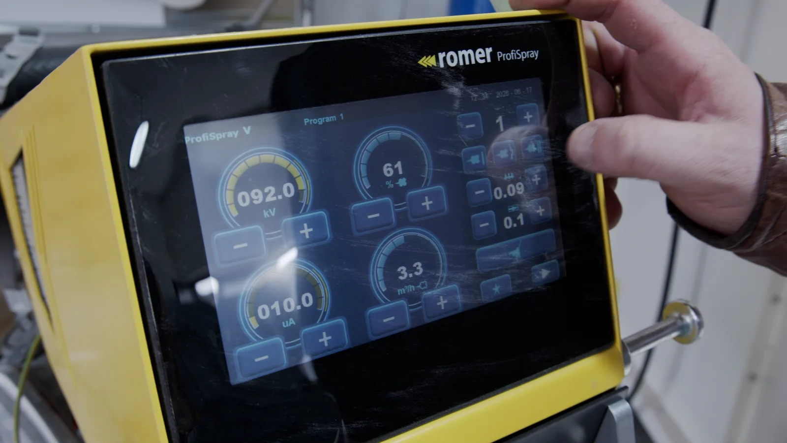 Ein gelbes Steuergeraet mit Touchscreen zeigt die Einstellungen fuer eine Pulverbeschichtungsanlage an, waehrend eine Hand es bedient.